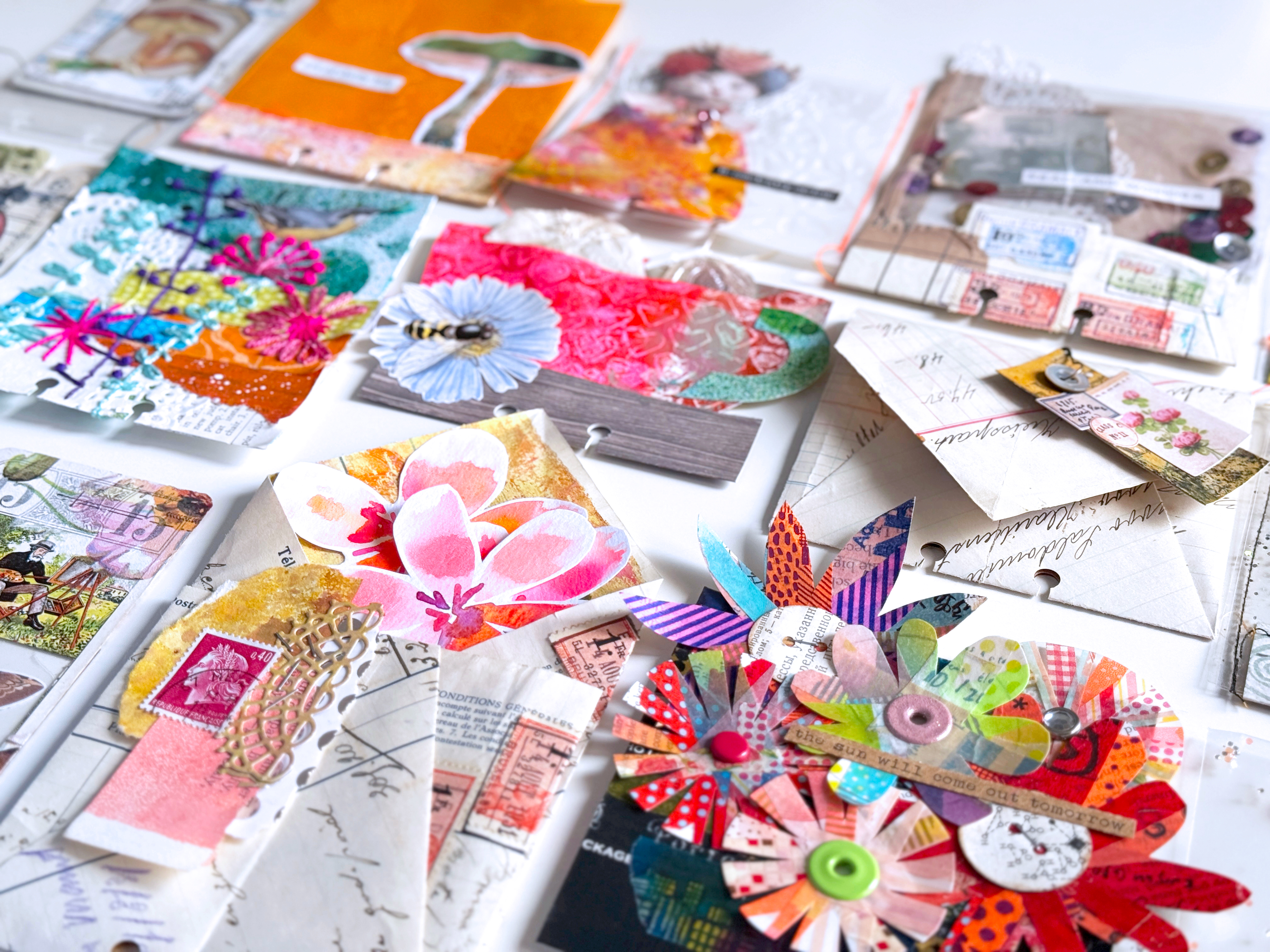 A colorful display of mixed media art pieces featuring layers of paper, flowers, textures, and various materials arranged on a white surface.